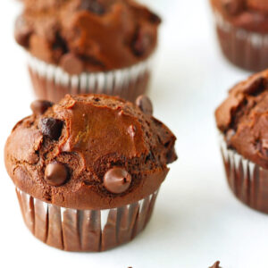 Close up of a Double Chocolate Chip Muffins on parchment paper with chocolate chips scattered in front. More muffins lined up behind.