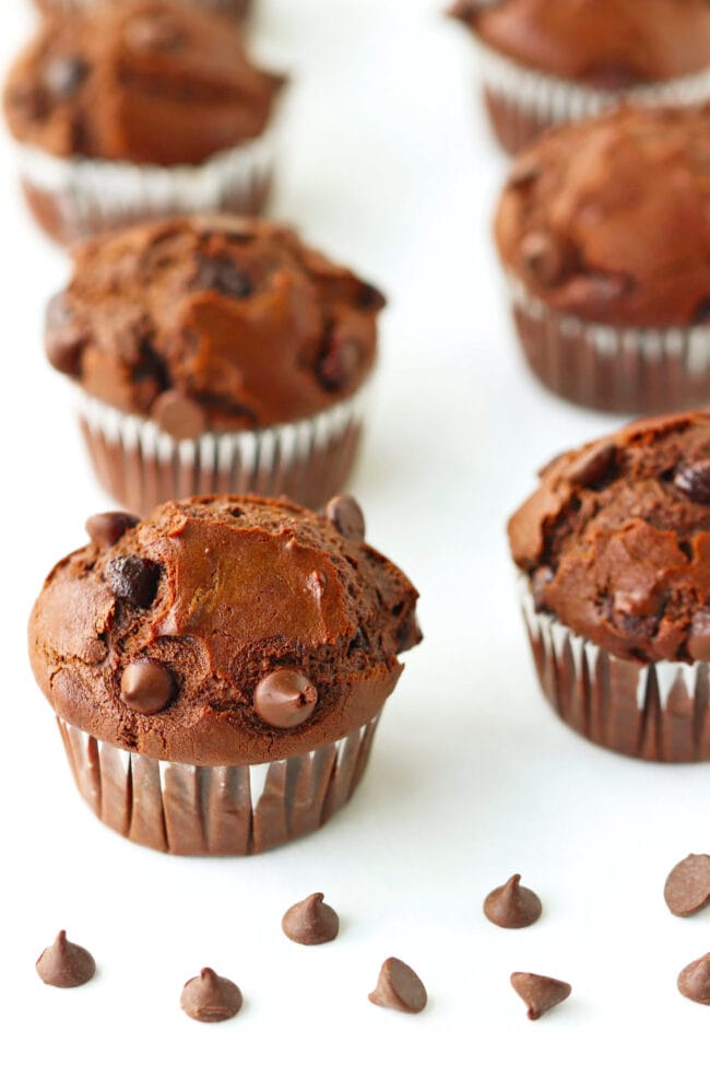 Double Chocolate Chip Muffins lined up on parchment paper and chocolate chips scattered in the front.
