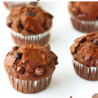 Muffins lined up on parchment paper and chocolate chips scattered in the front. Text overlay "Double Chocolate Chip Muffins".