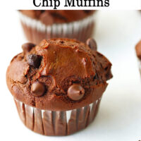Close up of a muffin on parchment paper and chocolate chips scattered in the front. Text overlay "Double Chocolate Chip Muffins".