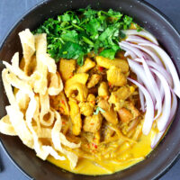 Top view of bowl with Khao Soi Gai with fried wonton wrapper strips, chopped coriander and mint leaves, and sliced red onion.
