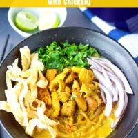 Front view of bowl with Khao Soi Gai topped with fried wonton wrapper strips, chopped coriander and mint leaves, and sliced red onion. Dutch oven with soup, and lime wedges and fried wonton wrapper strips in bowls behind. Text overlay "Khao Soi Gai (Northern Thai Coconut Curry Noodles Soup with Chicken)".