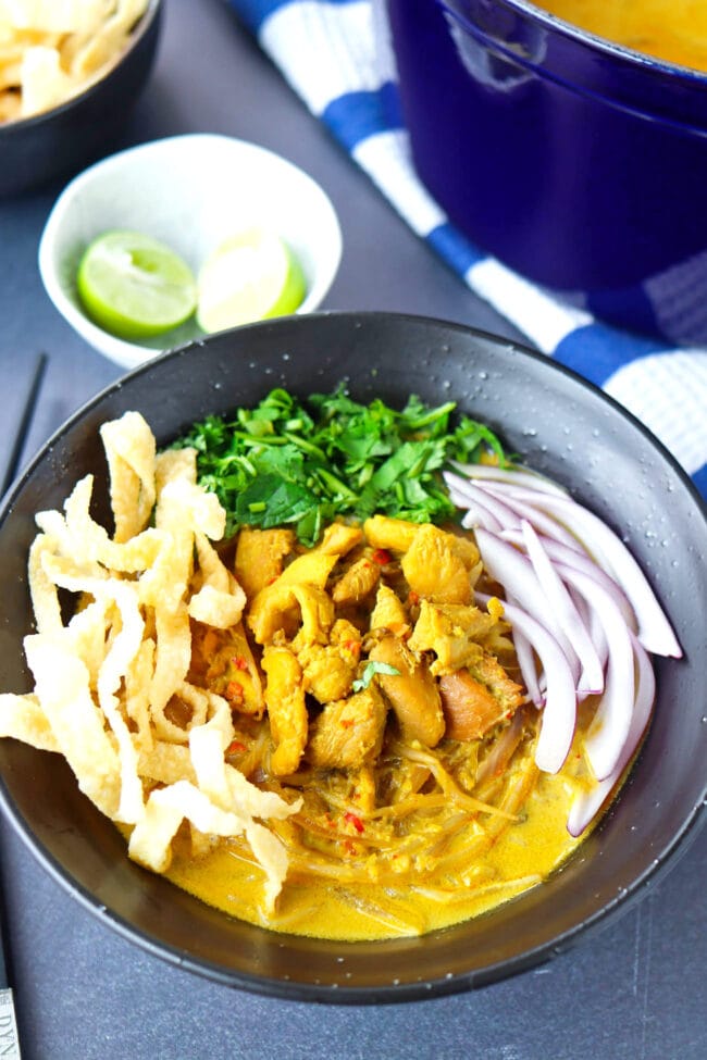Front view of bowl with Khao Soi Gai topped with fried wonton wrapper strips, chopped coriander and mint leaves, and sliced red onion. Dutch oven with soup, and lime wedges and fried wonton wrapper strips in bowls behind.