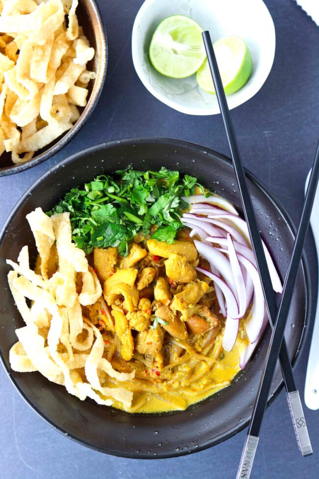 Top view of bowl with Khao Soi Gai with fried wonton wrapper strips, chopped coriander and mint leaves, and sliced red onion. Crossed chopsticks on top of side of the bowl, and lime wedges and crispy wonton wrapper strips in bowls behind.