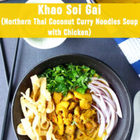 Top view of bowl with Khao Soi Gai topped with fried wonton wrapper strips, chopped coriander and mint leaves, and sliced red onion. Crossed chopsticks on and spoon on either side of bowl. Dutch oven with soup, and lime wedges and fried wonton wrapper strips in bowls behind. Text overlay "Khao Soi Gai (Northern Thai Coconut Curry Noodles Soup with Chicken)".