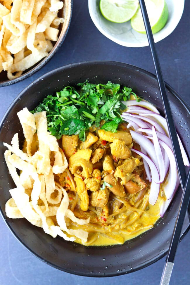 Close up top view of bowl with Khao Soi Gai with fried wonton wrapper strips, chopped coriander and mint leaves, and sliced red onion. Crossed chopsticks on top of side of the bowl, and lime wedges and crispy wonton wrapper strips in bowls behind.