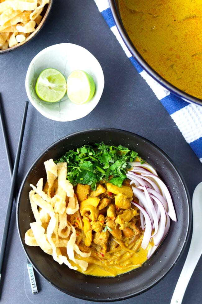 Top view of bowl with Khao Soi Gai topped with fried wonton wrapper strips, chopped coriander and mint leaves, and sliced red onion. Crossed chopsticks on and spoon on either side of bowl. Dutch oven with soup, and lime wedges and fried wonton wrapper strips in bowls behind.