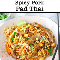 Front view of pork Pad Thai sprinkled with chopped peanuts on a plate with a lime wedge on the side. Wok with noodles and lime wedges in a small bowl in the back. Text overlay "Spicy Pork Pad Thai".