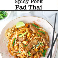 Top view of stir-fried thin rice noodles with seared pork slices on a plate with crushed peanuts, chopsticks, and a lime wedge. Wok with noodles, and small bowl with lime wedges in the back. Text overlay "Spicy Pork Pad Thai".