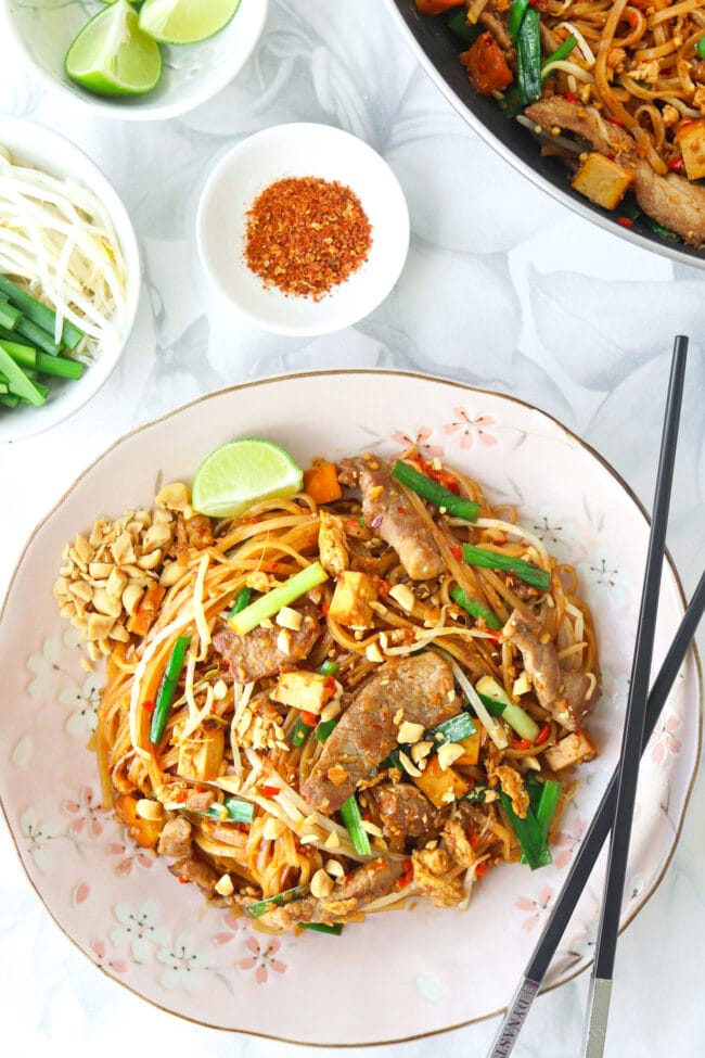 Top view of stir-fried thin rice noodles with seared pork slices on a plate with crushed peanuts, chopsticks, and a lime wedge. Wok with noodles, and small bowls with lime wedges, bean sprouts and Chinese chives, and ground hot pepper behind.