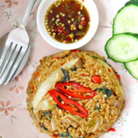 Top view of fried rice, cucumber slices, fish sauce with chopped chilies in small dish, and spoon and fork on a plate. Text overlay "Spicy Thai Basil Chicken Fried Rice".