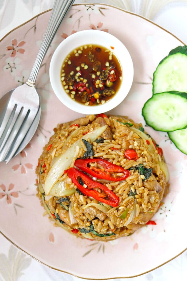 Closeup of fried rice, cucumber slices, fish sauce with chopped chilies, and spoon and fork on a plate.