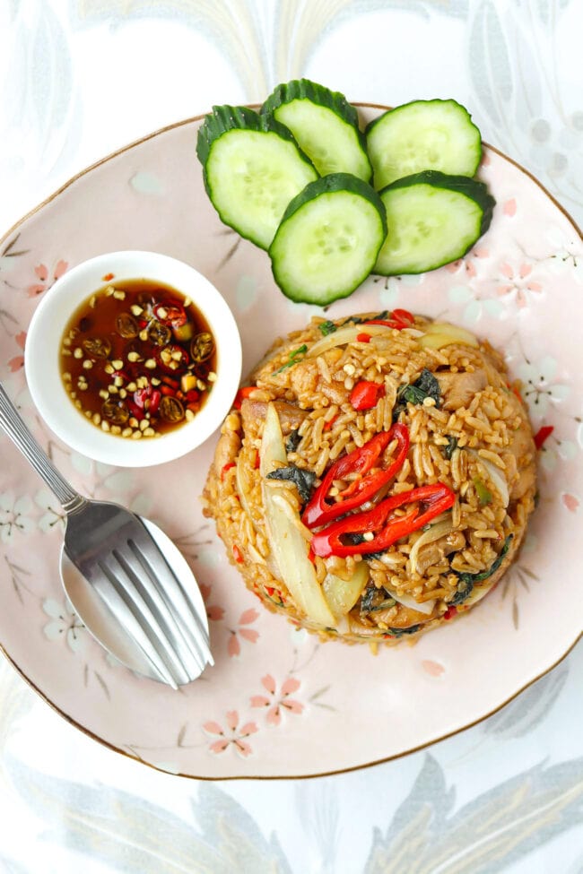 Top view of chicken fried rice on a plate with cucumber slices, fork and spoon, and small dish with chopped chilies in fish sauce.
