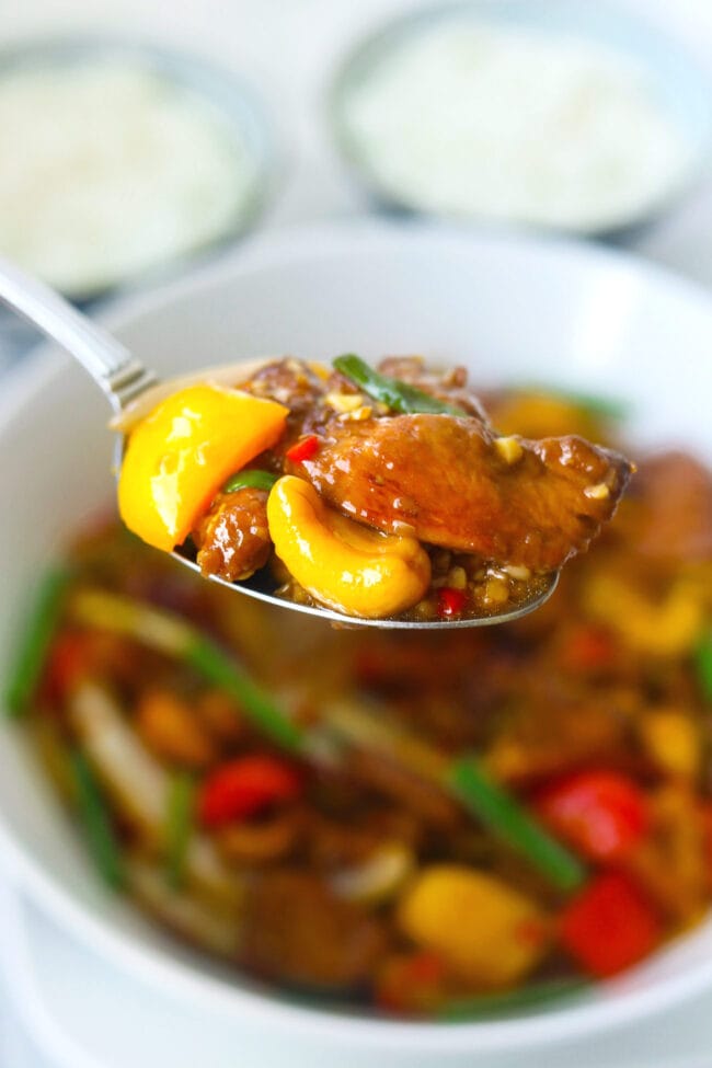 Spoon holding up chicken pieces, bell pepper, and a cashew nut above stir-fry dish.
