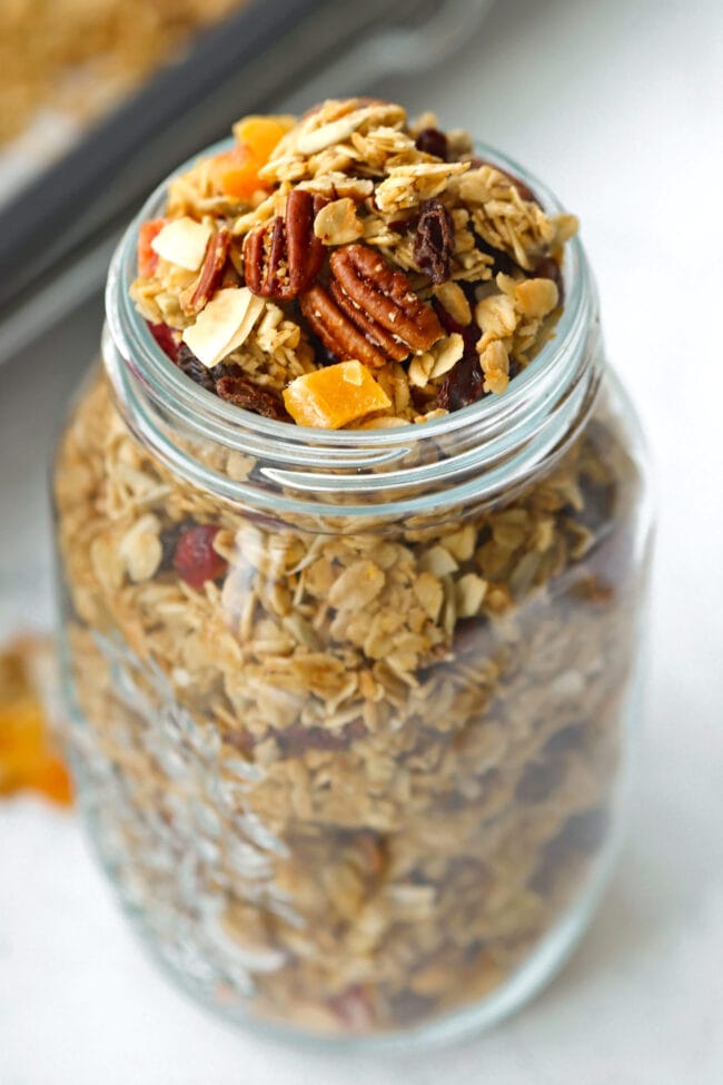 Tilted front view of granola in mason jar. Baking tray with granola in the back.