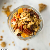 Top view of granola in mason jar surrounded by clusters on backdrop. Text overlay "Tropical Pecan Coconut Granola", "Easy", "Customizable", and "Refined Sugar Free".