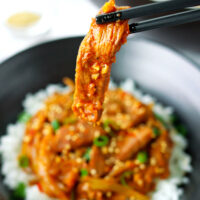 Chopsticks holding up a pork strip above bowl with pork stir-fry and rice. Text overlay "Spicy Korean Pork Stir-fry" and "thatspicychick.com".