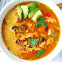 Close-up top view of white bowl of soup topped with diced avocado, cheese, and coriander. Text overlay "Spicy Pumpkin Chicken Tortilla Soup" and "thatspicychick.com".