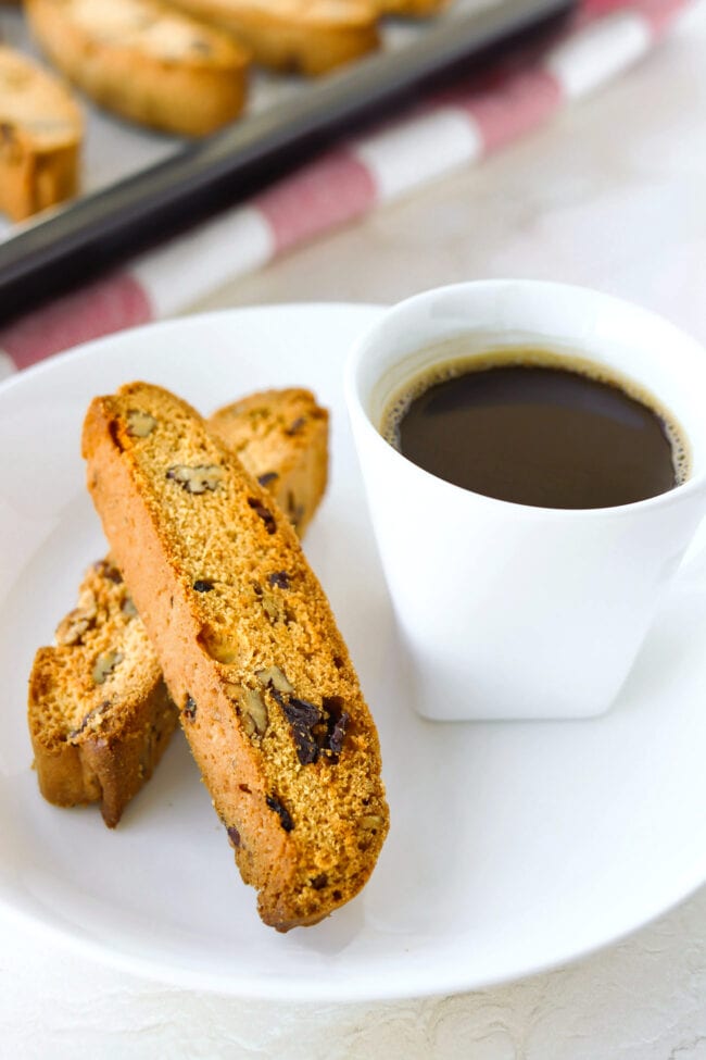 Close-up front view of two biscotti and a cup of coffee on a plate, and baking tray with biscotti in the back.