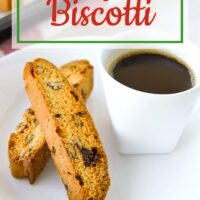 Close-up of two biscotti on a plate with a cup of coffee, and freshly baked biscotti on tray behind. Text overlay "Cranberry Pecan Biscotti" and "thatspicychick.com".