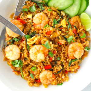 Top view of plate with spicy prawn fried rice on a plate with a spoon, fork, cucumber slices, and a lime wedge.