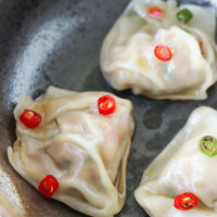 Close-up of three wontons in a black bowl topped with fish sauce and chopped chilies. Text overlay "Spicy Thai Basil Wontons, Pork, Chicken & Prawn Kra Pow Filling" and "thatspicychick.com".