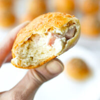 Fingers holding up a ham and cream cheese bagel ball with a bite taken out. Text overlay "Greek Yogurt Dough Stuffed Bagel Bombs" and "thatspicychick.com".