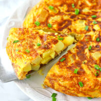 Front view of plate with a potato omelette and a wedge on a cake cutter. Text overlay "Spanish Omelette (Tortilla Española)", "thatspicychick.com", and "easy foolproof method".