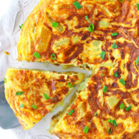 Top view of an omelette on a plate and a wedge on a cake cutter. Text overlay "Spanish Omelette (Tortilla Española)" and "thatspicychick.com".