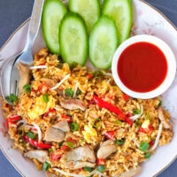 Close-up top view of pork fried rice on a plate with fork, spoon, cucumber, and Sriracha sauce.