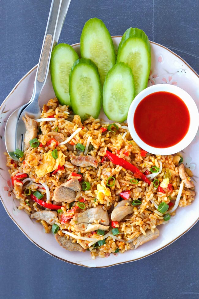 Top view of pork fried rice on a plate with fork, spoon, cucumber, and Sriracha sauce.