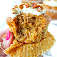 Hand holding up muffin with a bite out. Text overlay "Carrot Cake Muffins with Maple Cream Cheese Frosting" and "thatspicychick.com".