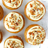 Top view of frosted muffins with chopped nuts on a plate. Text overlay "Carrot Cake Muffins with Maple Cream Cheese Frosting" and "thatspicychick.com".