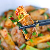 Chopsticks holding up a slice of tofu, pork, and garlic scape above serving bowl with stir-fry. Text overlay "Stir-fried Garlic Spaces with Pork & Tofu in Sichuan Sauce" and "thatspicychick.com".