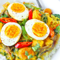 Close-up front view of egg curry on rice on a plate with a spoon. Text overlay "Thai Yellow Curry with Eggs & Everyday Veggies" and "thatspicychick.com".