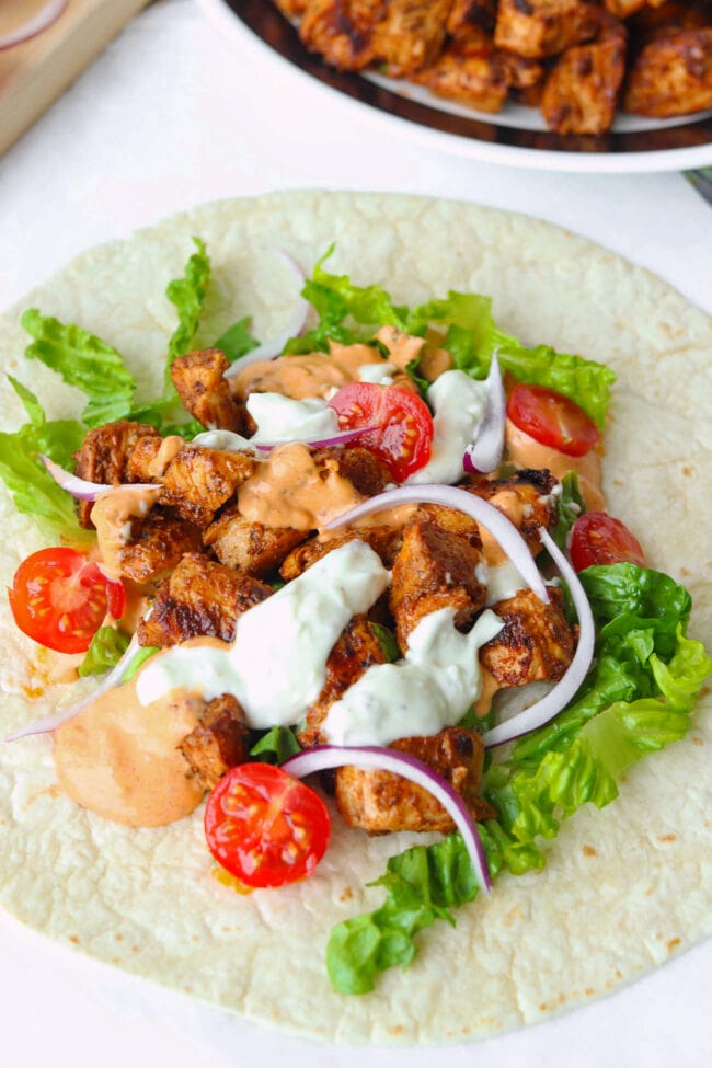 Close-up front view of chicken shish on tortilla with lettuce, cherry tomato halves, onion, and sauces.