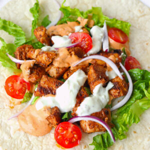 Close-up of chicken shish chunks, lettuce, sliced onion, cherry tomato halves, and sauces on a tortilla.