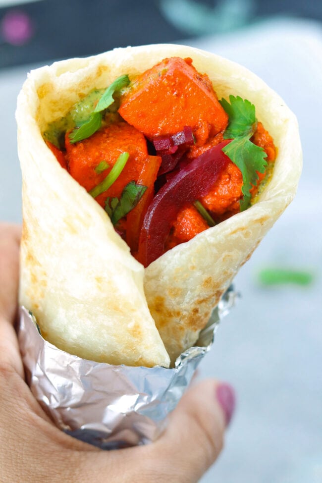 Hand holding up a paneer tikka kathi roll wrapped in foil.