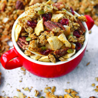 Granola in a red soup bowl. Text overlay "Apple Pie Granola", "Healthy | Vegan | Gluten-free", and "thatspicychick.com".