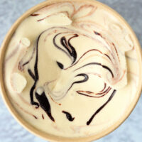 Top view of ice cream in a paper carton. Text overlay "Baileys Espresso Crème Ice Cream (No Churn)", "With Hot Fudge Sauce Swirl!", and "thatspicychick.com".