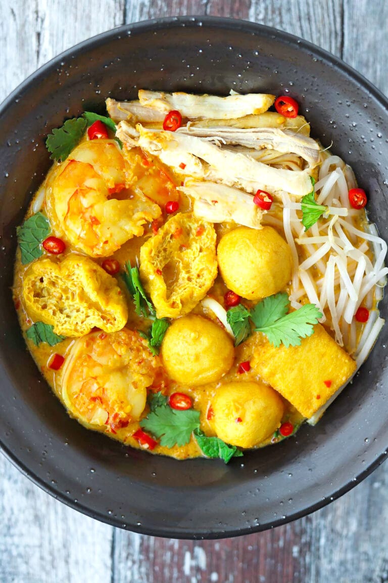 Closeup top view of spicy curry laksa in black bowl with garnishes.