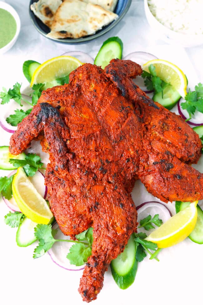 Tandoori chicken on a platter with red onion rings, cucumber slices, coriander sprigs and lemon wedges.