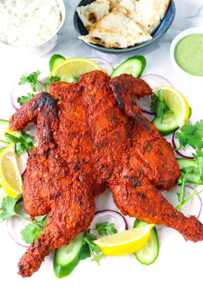 Whole Tandoori chicken on a serving platter. Rice, naan, and cilantro and mint sauce behind.