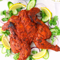 Tandoori chicken on a serving platter. Naan, rice, and cilantro and mint sauce in the back.