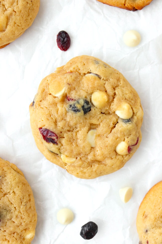 Close-up of cookie on parchment paper surrounded with dried berries and white chocolate chips.