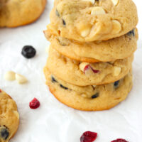 Close-up of stacked cookies on parchment paper surrounded by dried berries and white chocolate chips.