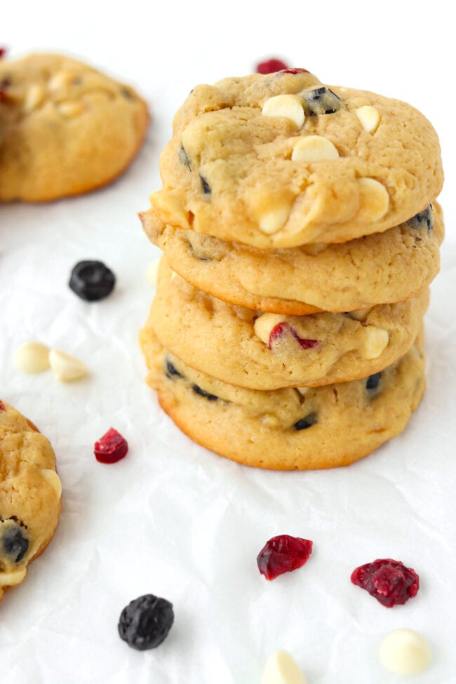 Close-up of stacked cookies on parchment paper surrounded by dried berries and white chocolate chips.