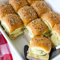 Front view of sliders in a baking and a slider on a spatula. Text overlay "Ham Egg and Cheese Breakfast Sliders" and "thatspicychick.com".