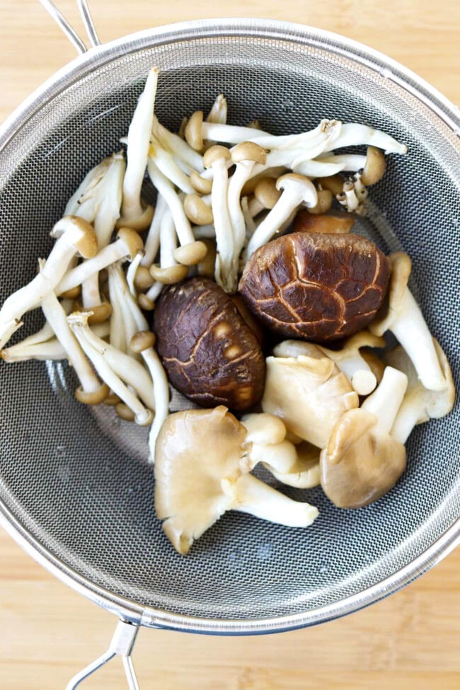 Assorted Asian mushrooms in a fine mesh strainer on top of a bowl.