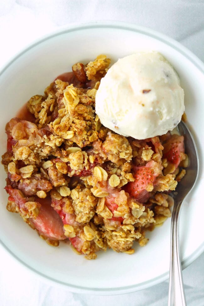 Strawberry crisp in bowl with a spoon and scoop of vanilla pecan ice cream.